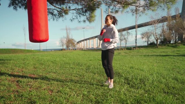Female boxer dreesd hoodie warming up legs muscule. Athletic female prepares for boxing workout outdor in park with punching bag. Fighter warm up body before training Female fitness. Self defence