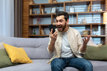 Mature man celebrating victory and successful achievement, got news online message on app using phone sitting on sofa in living room.