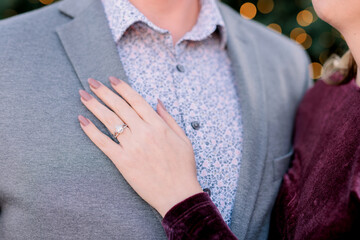 A woman's young hand is touching a young man wearing a flower blue dress shirt and gray blazer showing off her engagement ring.