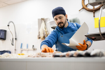 A middle-aged fishmonger in an apron and a hat is about to catch a fish from the counter, selling food, small and medium-sized business.