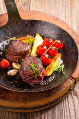 Traditional aged venison steak medallions with tomatoes, fries and mushrooms served as close-up in a rustic wrought-iron pan on a wooden board