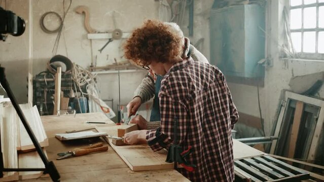 Elderly master and teen blogger influencer talking to camera recording tutorial video wood work course in carpentry.
