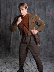 Fototapeta na wymiar A young guy in military-style clothes, a brown flight jacket and breeches with suspenders. Posing in the studio on a gray background