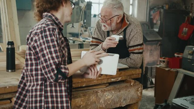 Old man and teenager drinking coffee and laughing using tablet during break in carpentry home workshop