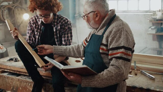 Teen boy in a carpentry workshop sitting on the table with tools, making notes with his grandfather making the new bass guitar