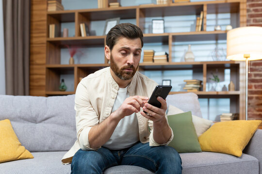 A Man Sitting On A Couch At Home, Looking Bored As He Scrolls Through Dating Apps On His Phone. The Casual Setting And Relaxed Posture Create A Relatable Image Of Modern Dating Culture