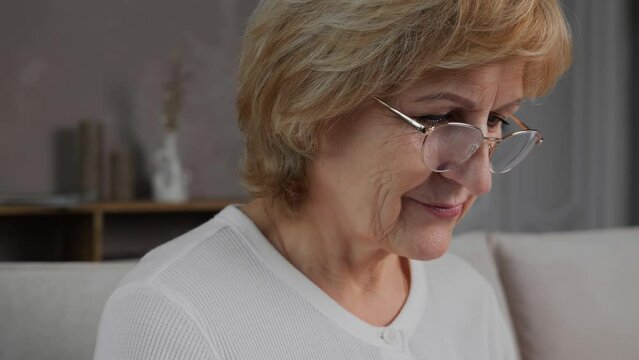 Close-up of the face of an adult 60s female blogger or freelancer who works on a laptop while sitting at home. A close-up portrait of a modern cute elderly woman with glasses working on a computer. 4K
