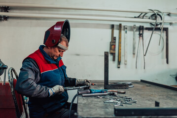 Close up welder in protective uniform and mask welding metal pipe on the industrial table with other workers behind in the industrial workshop.