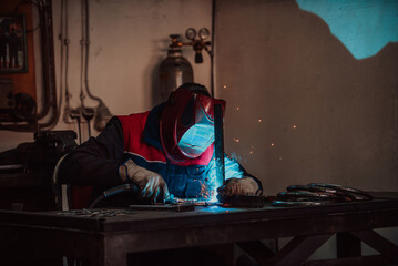 Fototapeta na wymiar Profesional welder in protective uniform and mask welding metal pipe on the industrial table with other workers behind in the industrial workshop.