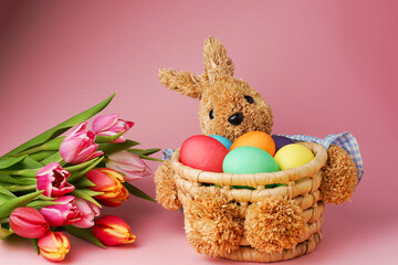 Straw bunny basket with easter colorful eggs und fresh tulips on pink background. Happy Easter card. Close up