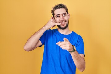 Hispanic man with beard standing over yellow background smiling doing talking on the telephone gesture and pointing to you. call me.