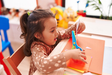 Adorable hispanic girl student sitting on table cutting paper at kindergarten