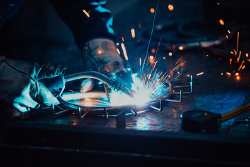 Close up welder in protective uniform and mask welding metal pipe on the industrial table with other workers behind in the industrial workshop.