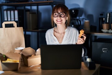 Young beautiful woman working using computer laptop and eating delivery food smiling looking to the side and staring away thinking.