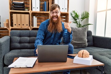Redhead man with long beard using laptop working at psychology clinic pointing thumb up to the side...