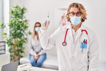 Blond man wearing doctor uniform and medical mask holding syringe scared and amazed with open mouth for surprise, disbelief face