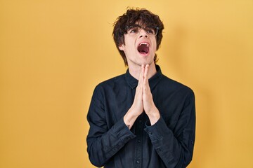 Young man wearing glasses over yellow background begging and praying with hands together with hope expression on face very emotional and worried. begging.