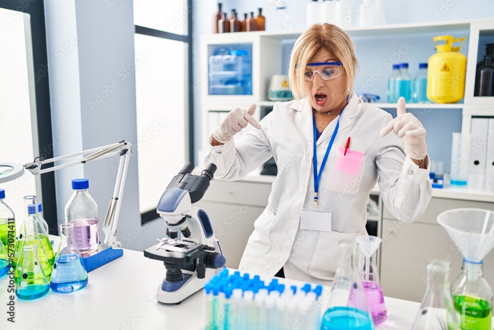 Wall mural middle age blonde woman working at scientist laboratory pointing down with fingers showing advertise