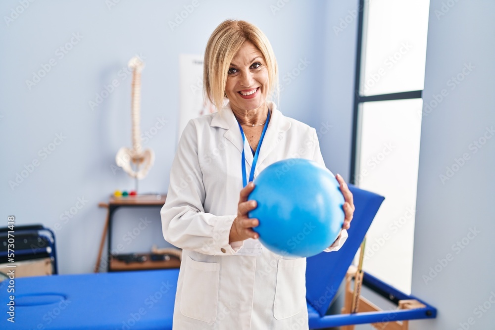 Sticker middle age blonde woman wearing physiotherapist uniform holding ball at physiotherapy clinic