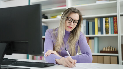 Young blonde woman student using computer writing on document at library university