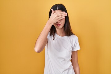 Young beautiful woman standing over yellow background covering eyes with hand, looking serious and sad. sightless, hiding and rejection concept