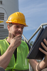 Male engineer with short brown hair, wearing yellow hard hat and reflective vest, inspecting wind turbine using digital tablet, thoughtful