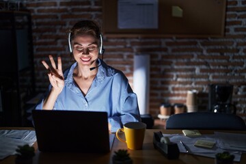 Beautiful brunette woman working at the office at night showing and pointing up with fingers number three while smiling confident and happy.