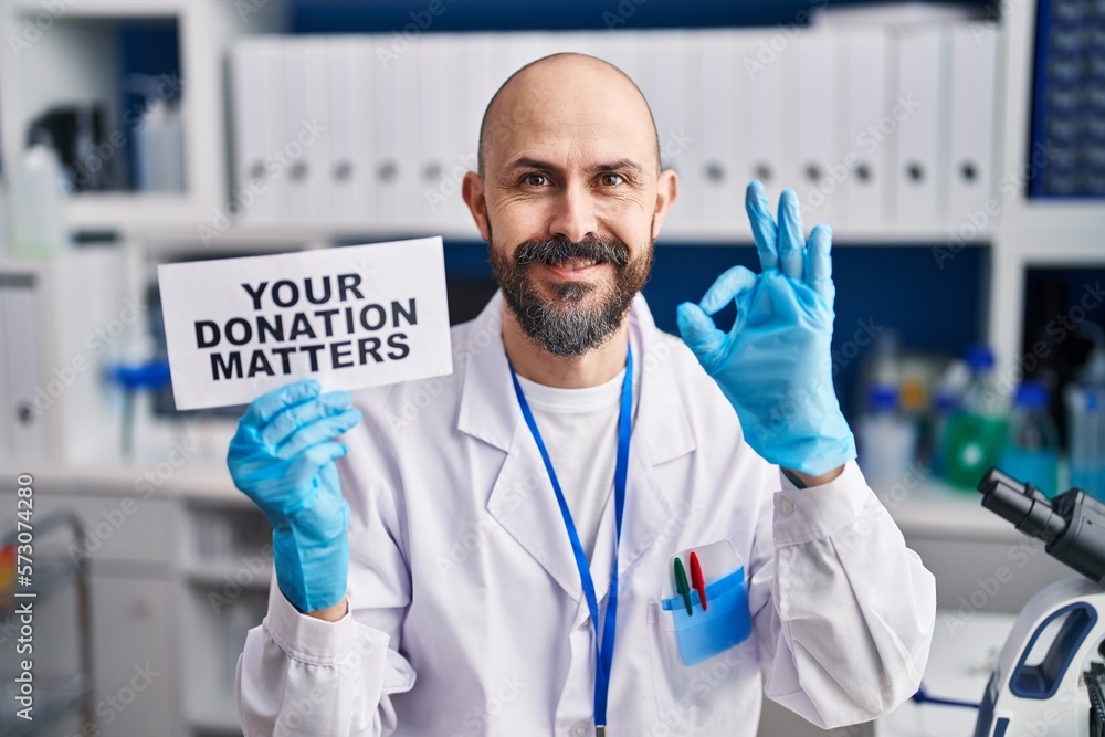 Sticker Young hispanic man working at scientist laboratory holding you donation matters banner doing ok sign with fingers, smiling friendly gesturing excellent symbol