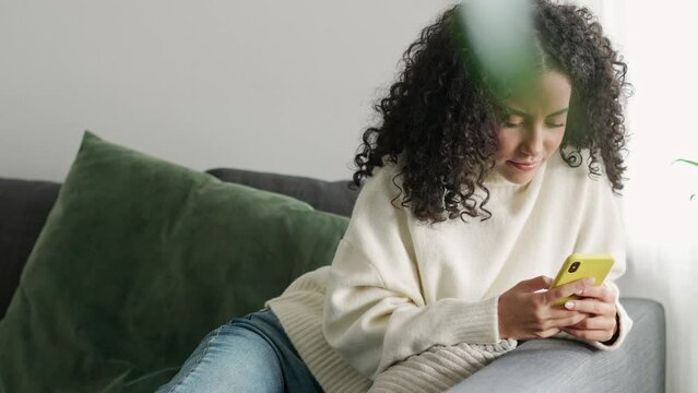 Young pretty latin woman sitting on couch using cell phone, holding smartphone, looking at cellphone doing online shopping in mobile digital shop app, checking social media commercial offers.