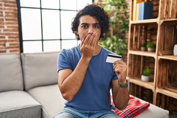 Hispanic man with curly hair holding credit card covering mouth with hand, shocked and afraid for mistake. surprised expression