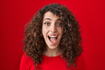 Hispanic woman with curly hair standing over red background celebrating crazy and amazed for success with open eyes screaming excited.