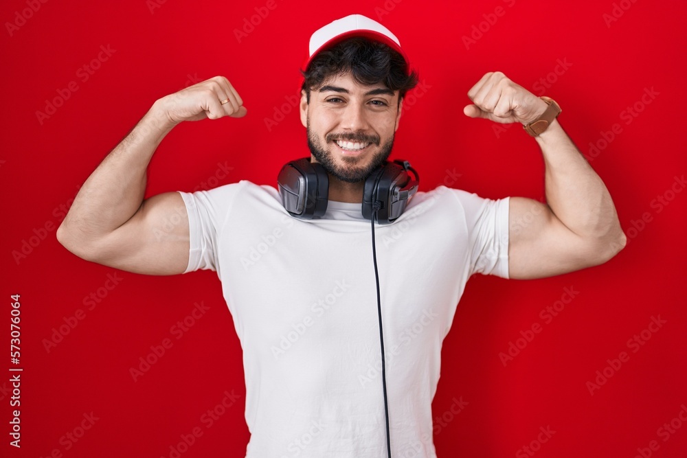 Wall mural hispanic man with beard wearing gamer hat and headphones showing arms muscles smiling proud. fitness