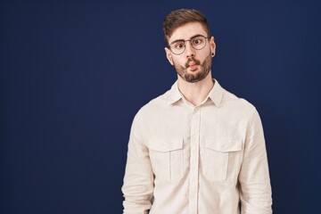 Hispanic man with beard standing over blue background making fish face with lips, crazy and comical gesture. funny expression.