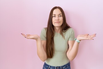 Beautiful brunette woman standing over pink background clueless and confused expression with arms...