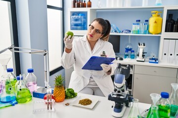 Young beautiful plus size woman scientist holding apple reading document at pharmacy