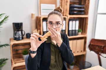 Young hispanic girl eating protein bar as healthy energy snack at the office covering mouth with...