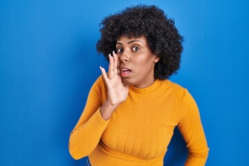 Fototapeta na wymiar Black woman with curly hair standing over blue background hand on mouth telling secret rumor, whispering malicious talk conversation