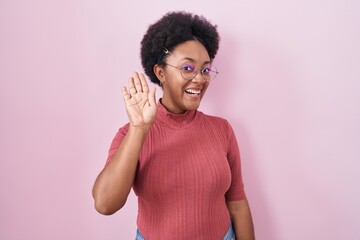 Beautiful african woman with curly hair standing over pink background waiving saying hello happy and smiling, friendly welcome gesture