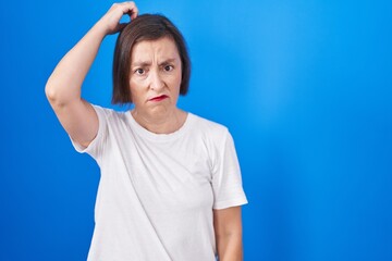 Middle age hispanic woman standing over blue background confuse and wondering about question. uncertain with doubt, thinking with hand on head. pensive concept.