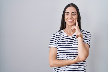 Young brunette woman wearing striped t shirt looking confident at the camera smiling with crossed arms and hand raised on chin. thinking positive.