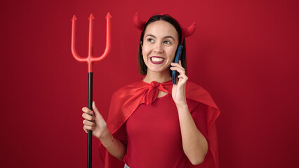 Young beautiful hispanic woman wearing devil costume talking on smartphone over isolated red background