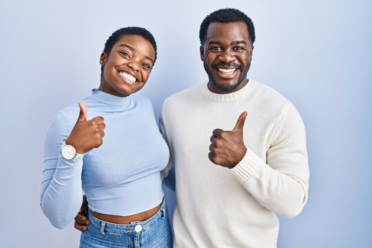 Young african american couple standing over blue background doing happy thumbs up gesture with hand. approving expression looking at the camera showing success.