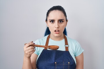 Young modern girl with blue hair wearing cook apron holding spoon scared and amazed with open mouth for surprise, disbelief face