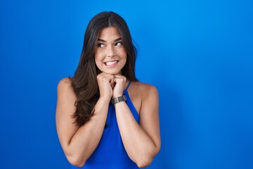 Hispanic woman standing over blue background laughing nervous and excited with hands on chin...