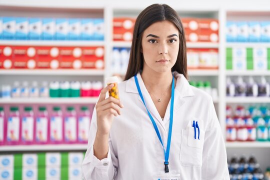 Hispanic woman working at pharmacy drugstore holding pills thinking attitude and sober expression looking self confident