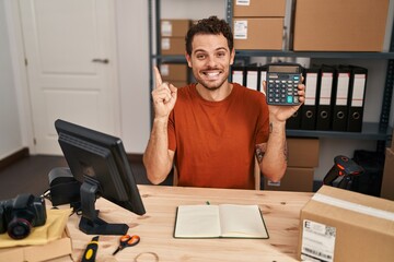 Young hispanic man working at small business ecommerce holding calculator smiling happy pointing with hand and finger to the side