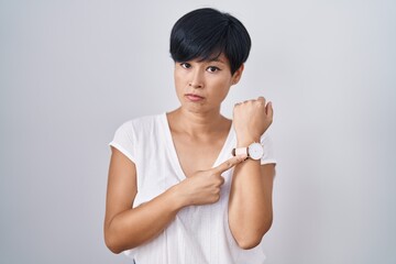 Young asian woman with short hair standing over isolated background in hurry pointing to watch...