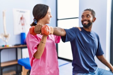 Fototapeta na wymiar Man and woman wearing physiotherapist uniform having rehab session stretching arm holding dumbbell at physiotherpy clinic