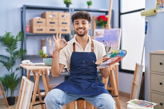 Arab man with beard painter sitting at art studio holding palette doing ok sign with fingers, smiling friendly gesturing excellent symbol