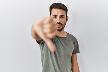 Young hispanic man with beard wearing casual t shirt over white background looking unhappy and angry showing rejection and negative with thumbs down gesture. bad expression.